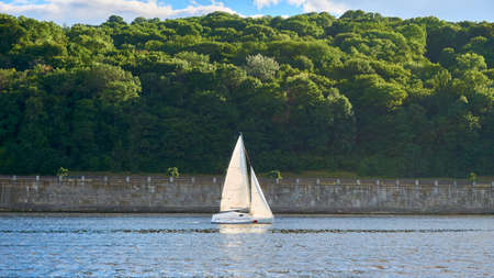 Small yacht on the background of the coast with a stone wall and forestの写真素材