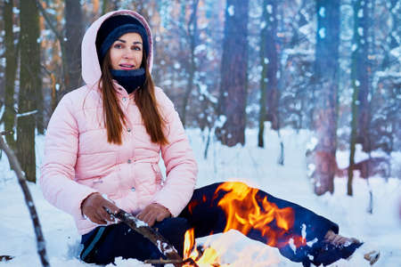 Young woman sitting near the fire in the winter forest