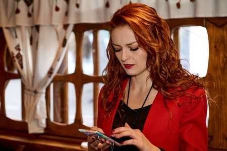 Serious pretty red-haired girl in a red jacket writes a message on the phone