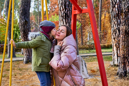 Cute child gently kisses and hugs her mom on a swing in the autumn forest park