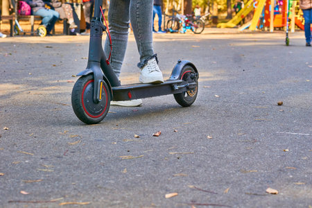 Female legs in gray jeans on an electric scooter in a public park