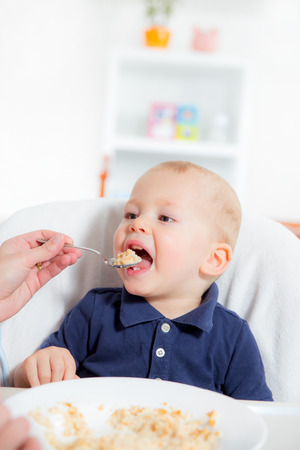Mother feeding hungry baby in the highchair indoorsの写真素材