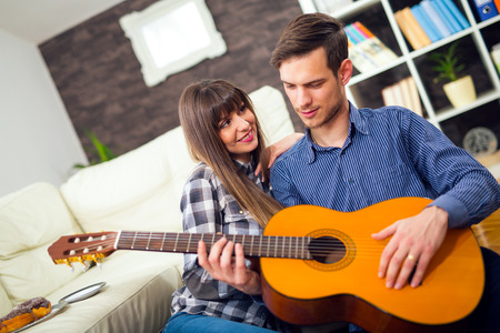 Young and attractive couple sitting near the couch with a guitar. The male is playing the guitar. Selective focusの写真素材
