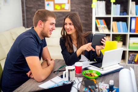 Beautiful smiling young businesswoman sitting working with a young man in a busy office as they discuss information on a digital tabletの写真素材