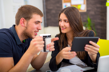 Beautiful smiling young businesswoman sitting working with a young man in a busy office as they discuss information on a digital tabletの写真素材