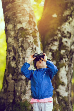 Young boy in a blue coat looking through binoculars in the forestの写真素材