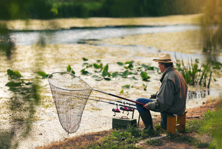 Senior man fishing on a freshwater lake sitting patiently on the shore with his rod and reel as he enjoys his retirementの写真素材