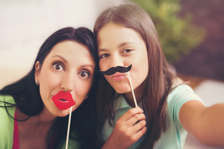 Mother taking selfie with daughter holding artificial mustache and lips stick at homeの写真素材