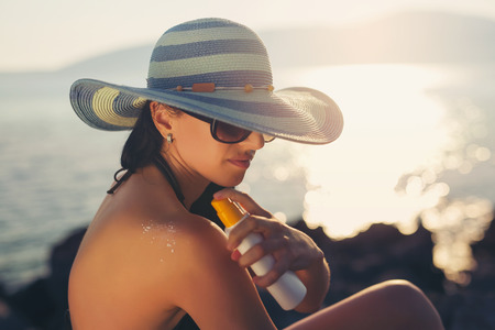 Young woman in sunglasses holding bottle of sunscreen lotion, spraying sunblock cream on shoulder...