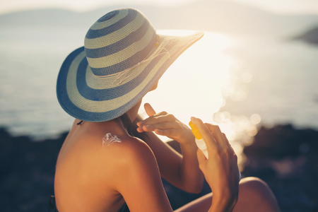 Young woman in sunglasses holding bottle of sunscreen lotion, spraying sunblock cream on shoulder before tanningの写真素材