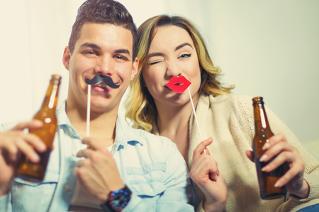 Young couple having fun holding artificial mustache and lips stick and beerの写真素材