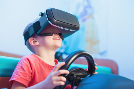 Happy little boy in virtual reality glasses playing video game with racing wheels at homeの写真素材