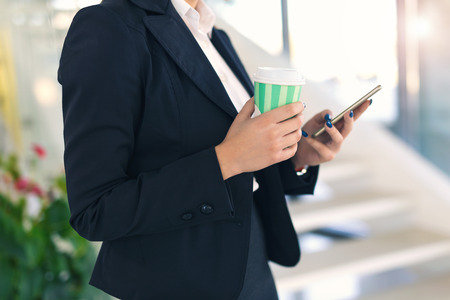Close up of young businesswoman drinking coffee or milkshake while holding a phoneの写真素材