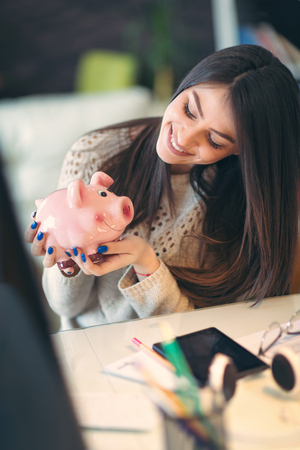 Business woman sitting at a desk and holding piggy bankの写真素材