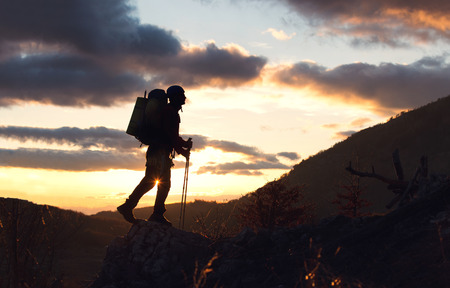 Young mountaineer standing with backpack on top of a mountain silhouette in mountains with sunset...