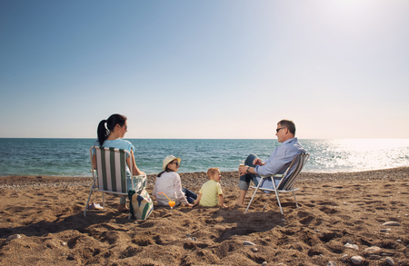 Portrait of a happy family in summer nature. Mother and father are sitting on a beach deck chair, daughter and son playingの写真素材