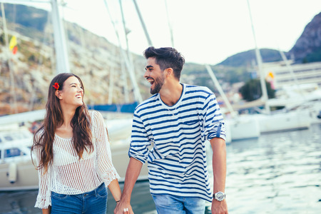 Attractive couple exploring on their summer vacation laughing as they hold hands while sightseeing a small boat harbour on the coastの写真素材