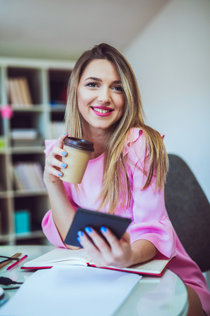 Time for coffee break. Young woman in smart casual wear holding a cup of coffee and looking at camera while sitting at her working place in creative officeの写真素材