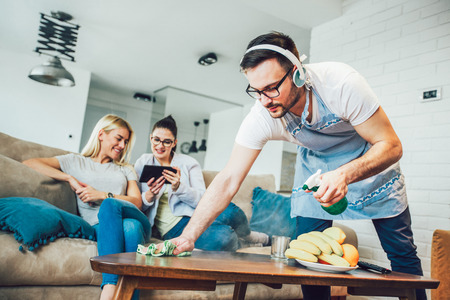 Husband housekeeping and cleaning concept. A man cleans the house, while women gossiping on the sofaの写真素材