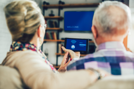 Elderly couple sitting comfortably on a sofa with their backs holding remote home control system on a digital tabletの写真素材