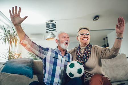 Happy senior couple watching soccer on tv and celebrating victory at homeの写真素材