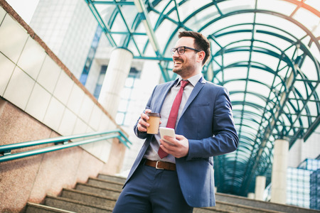 Businessman holding mobile phone and cup of coffee outside of office buildings in the background. の写真素材
