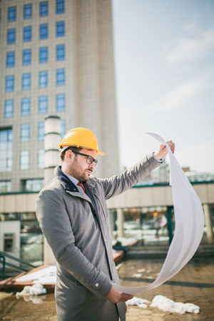 Architect in protective helmet holding blueprint in front of office buildingの写真素材