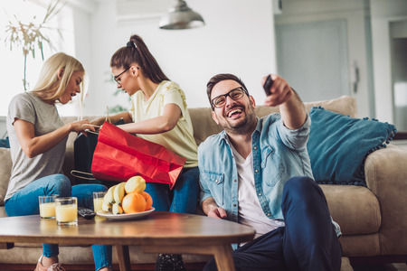 Young women sitting at home with bags after shopping. Young man watching tv.の写真素材