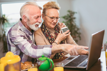 Excited senior couple looking at a laptop together in the kitchen pointing and smiling at something amusing on the screenの写真素材