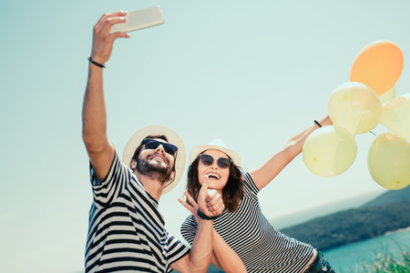 Smiling couple wearing sunglasses with balloons make selfie photo over sea backgroundの写真素材