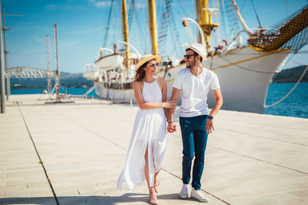 Happy young couple walking by the harbor of a touristic sea resort with sailboats on backgroundの写真素材