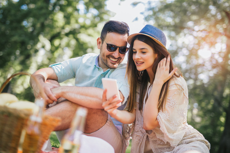 Couple lying on a picnic blanket in a park with a picnic basket filled with fruit, they are using smart phoneの写真素材