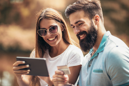 Cheerful young couple sitting on a park bench and uses a digital tablet for online shoppingの写真素材