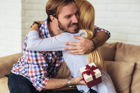 Smiling young man surprising cheerful woman with a gift box at homeの写真素材
