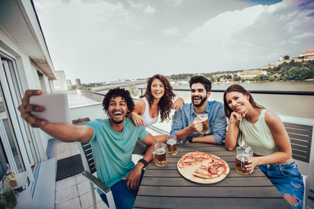 Friends enjoying pizza. Group of young cheerful people eating pizza and drinking beer while sitting at the bean bags on the roof of the buildingの写真素材