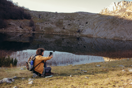 Thoughtful young man studying map on modern digital tablet on nature while sitting near lakeの写真素材
