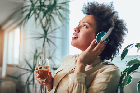 Young beautiful african american woman relaxing and listening to music using headphone, drinking wine.の写真素材