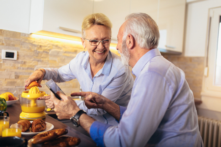 Aged couple busy look at digital tablet while having delicious breakfast at home kitchenの写真素材