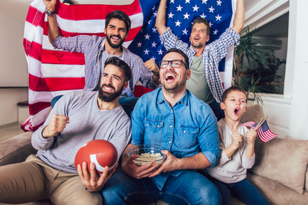 Male friends watching tv and cheering sport games on sofa at home. Group of friends watching american football match.の写真素材