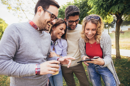 Four happy smiling young friends walking outdoors in the park holding digital tablet