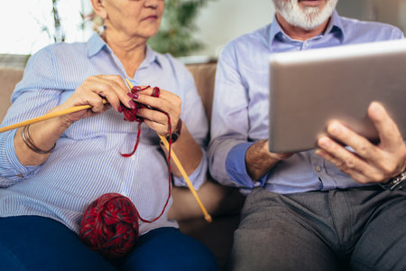 Senior woman knitting woollen clothes and her husband is using digital tablet.