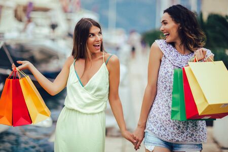 Female friends with shopping bags walking by the harbor of a touristic sea resort with boats on backgroundの写真素材