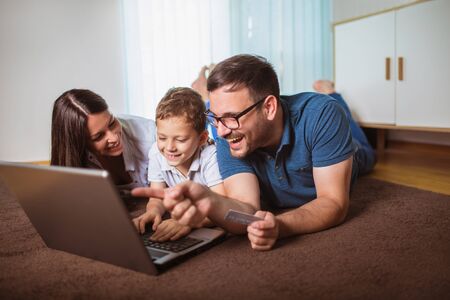 Beautiful parents and their son are doing shopping online using laptop and smiling at home.の写真素材