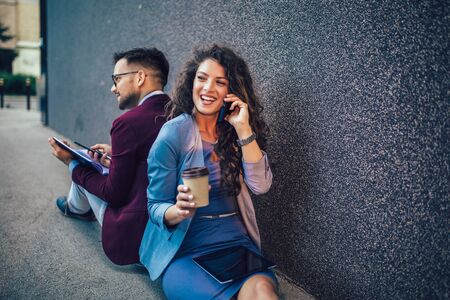 Young entrepreneurs sitting down in front of a modern office building and using phone. Brainstorming outside in a beautiful day.の写真素材