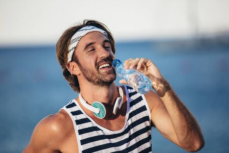 Portrait of healthy athletic man with fit body holding bottle of refreshing water, resting after workout or running at beach.の写真素材