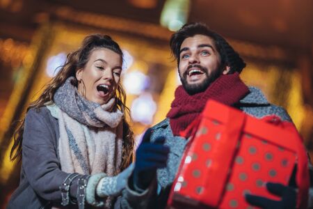 Young couple in the city centre with holiday's brights in background. Man presenting gift to woman.の写真素材