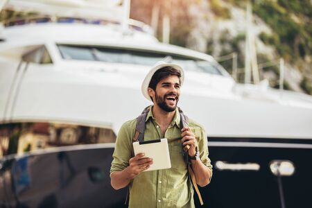 Young man walking by the harbor of a touristic sea resort with boats on background, holding digital tablet.の写真素材