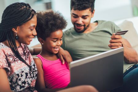 Cute little Afro-American girl and her beautiful young parents using a laptop and doing shopping online while sitting on a sofa at home.の写真素材