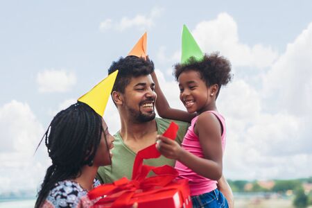 Happy black family at home. African american father, mother and child celebrating birthday, having fun at party. Young woman giving gift to daughterの写真素材