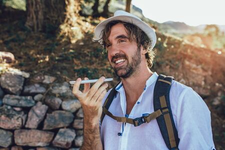 Fashionable man tourist using voice command recorder on smartphone in forest.